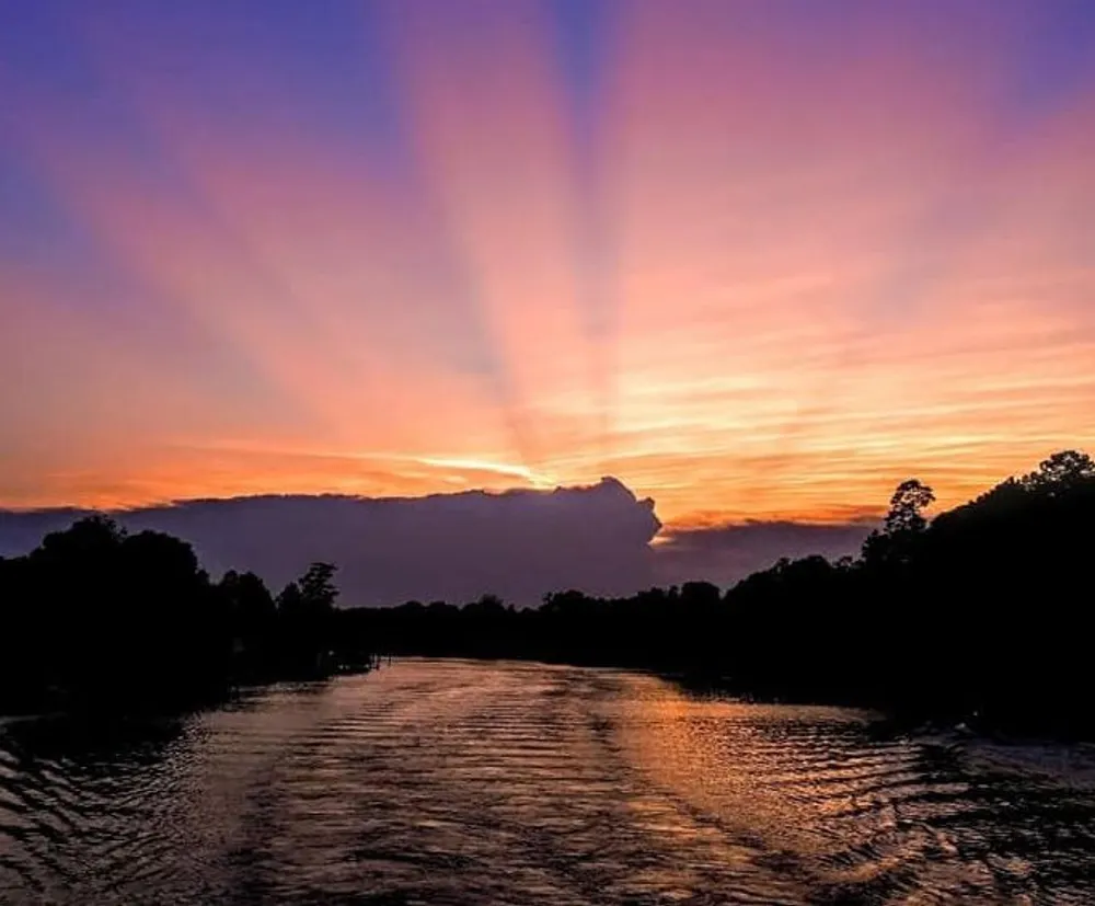 The image captures a picturesque sunset with vibrant rays of light casting a warm glow over a serene river bordered by silhouetted trees