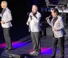 Three men in suits are performing on stage singing into microphones with stage lighting in the background