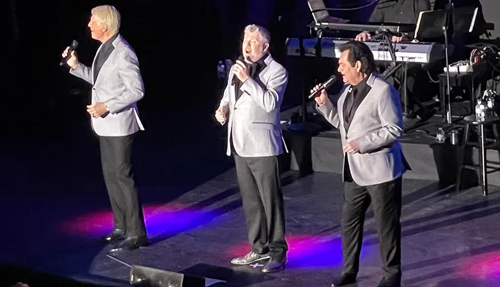 Three men in matching silver jackets are performing on stage with microphones in hand against a backdrop of musical instruments