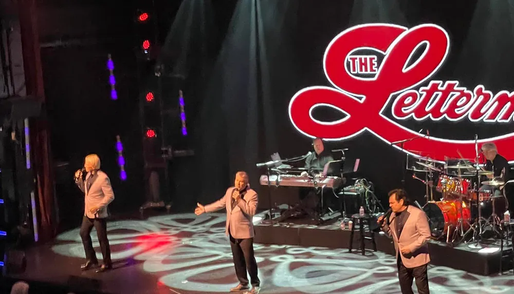 Four performers likely part of a vocal group named The Lettermen based on the backdrop are on stage during a live performance with musicians in the background