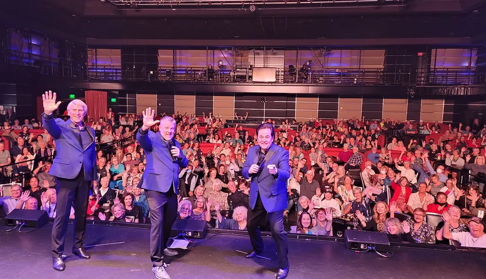 Three performers on stage are waving to an enthusiastic audience in a well-lit venue