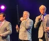 Three men in suits are performing on stage singing into microphones with stage lighting in the background
