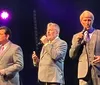 Three men in suits are performing on stage singing into microphones with stage lighting in the background