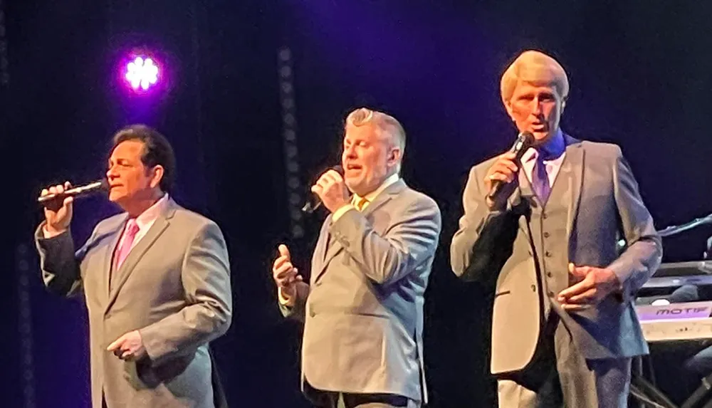 Three men in suits are performing on stage singing into microphones with stage lighting in the background