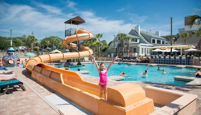 A cheerful child stands with arms raised in front of a winding waterslide at a busy outdoor pool on a sunny day