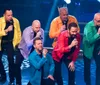 A group of six men are energetically performing on stage singing into microphones with dynamic expressions and colorful jackets