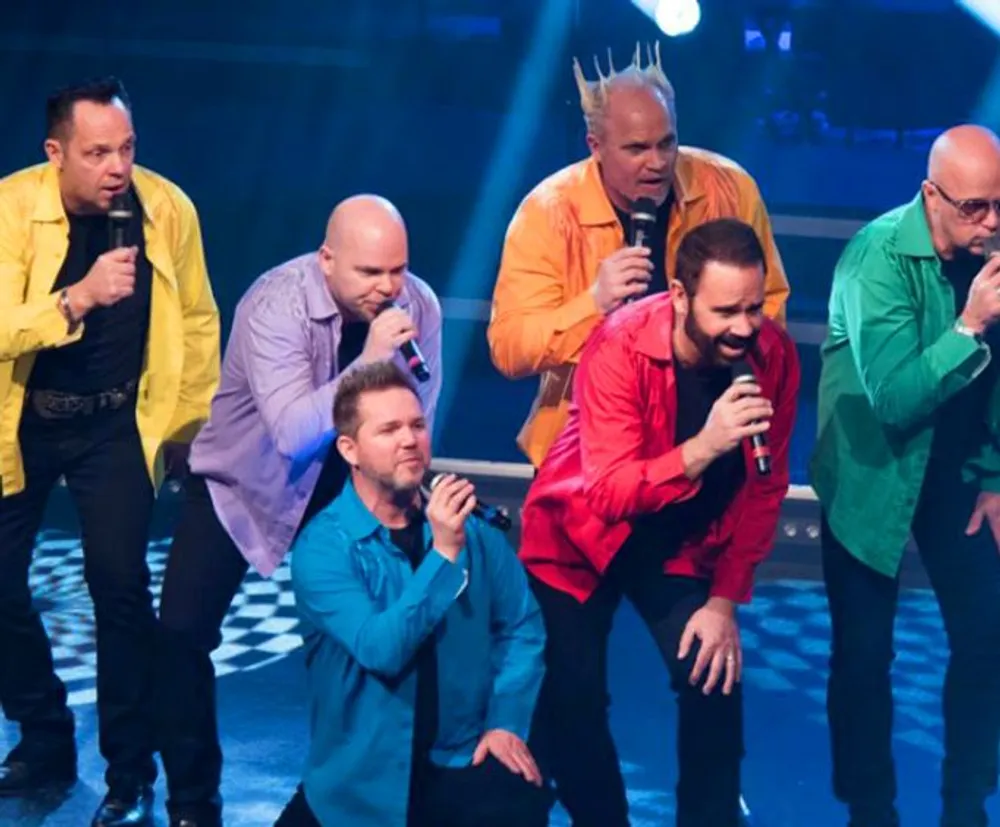 A group of six men are singing on stage each wearing a different vibrant-colored shirt with some holding microphones