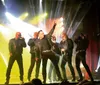 A group of six men are energetically performing on stage singing into microphones with dynamic expressions and colorful jackets