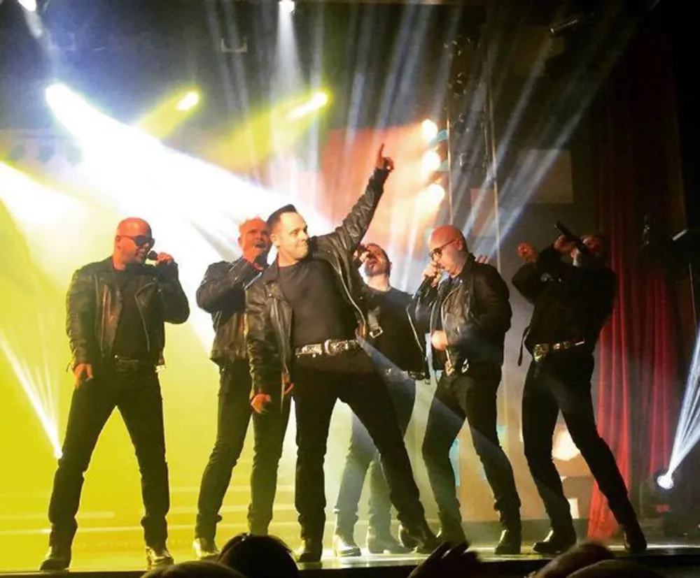 A group of performers in matching black outfits and sunglasses is energetically engaging with an audience on a brightly lit stage