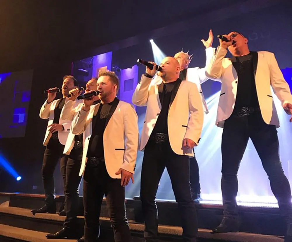 Five men are dressed in white jackets and black pants performing on stage with microphones in hand