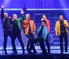 A group of six men are energetically performing on stage singing into microphones with dynamic expressions and colorful jackets