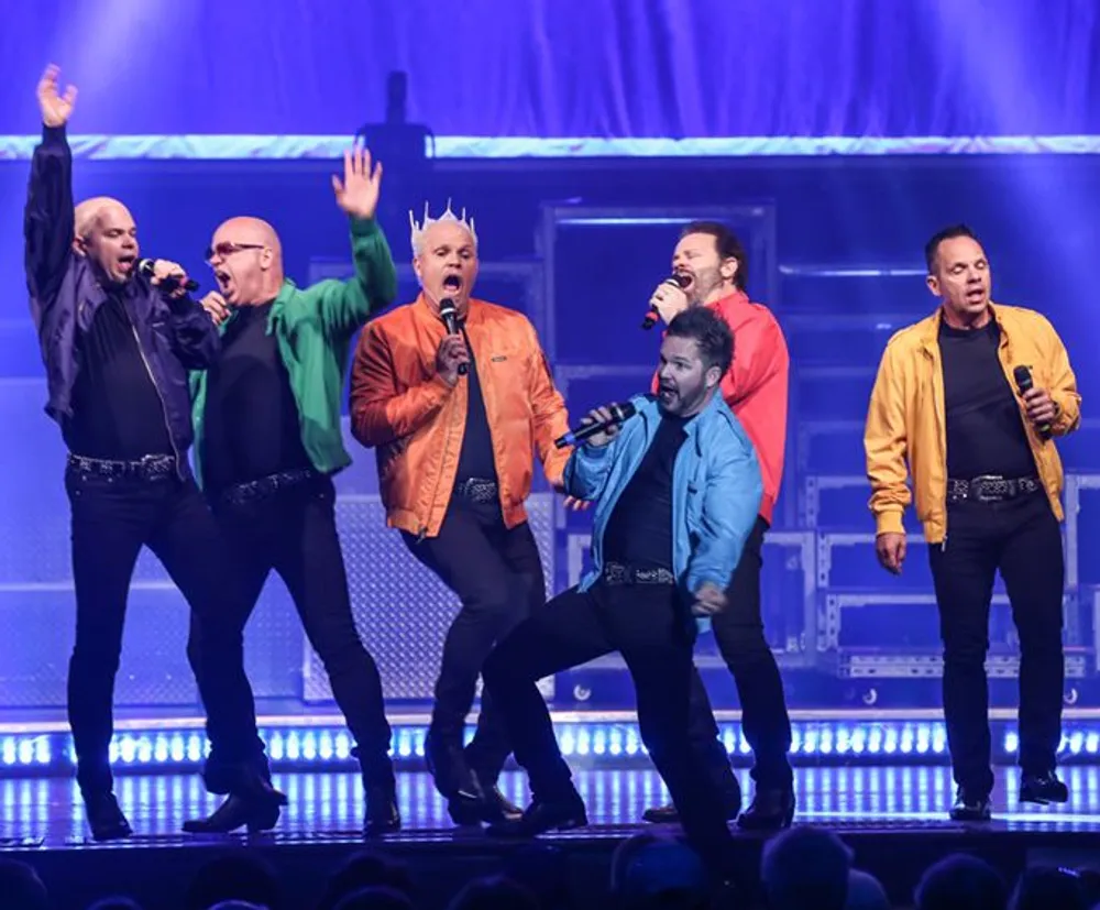 A group of six men are energetically performing on stage singing into microphones with dynamic expressions and colorful jackets