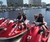 Four people are sitting on red jet skis at a marina on a sunny day