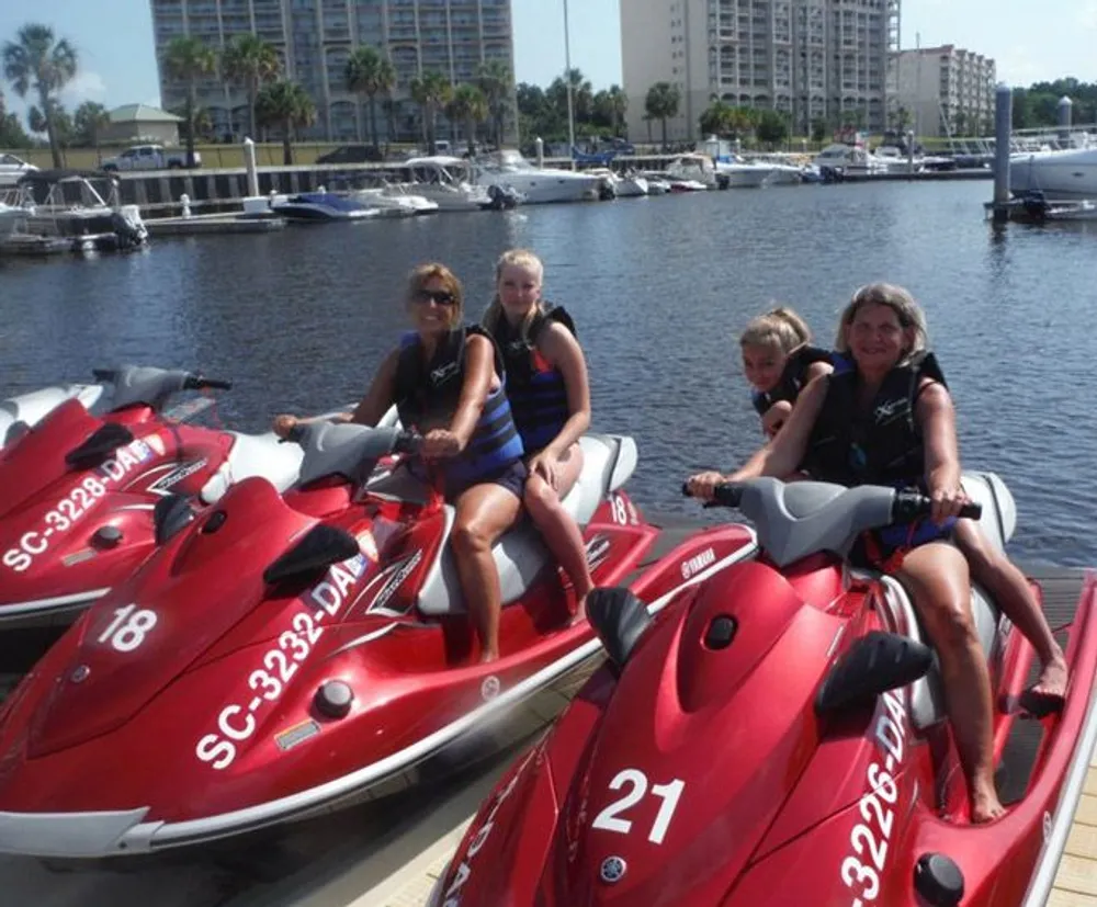 Four people are sitting on red jet skis at a marina on a sunny day