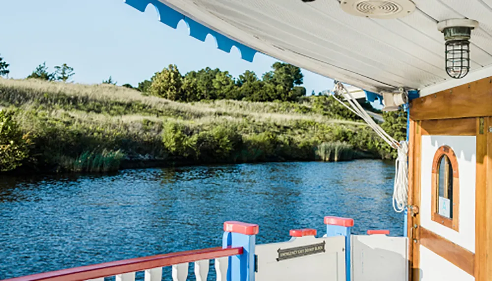 The image shows a scenic view from the deck of a boat highlighting the river and the lush greenery on the riverbank under a clear sky