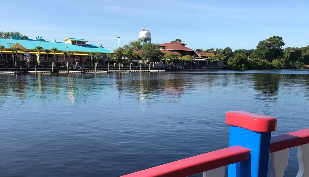 The image shows a calm waterway with colorful waterfront buildings a water tower in the background and a railing with red and blue details in the foreground