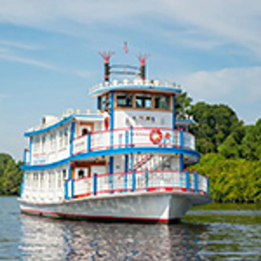 A white and blue riverboat with red accents is cruising on a calm river amidst lush greenery