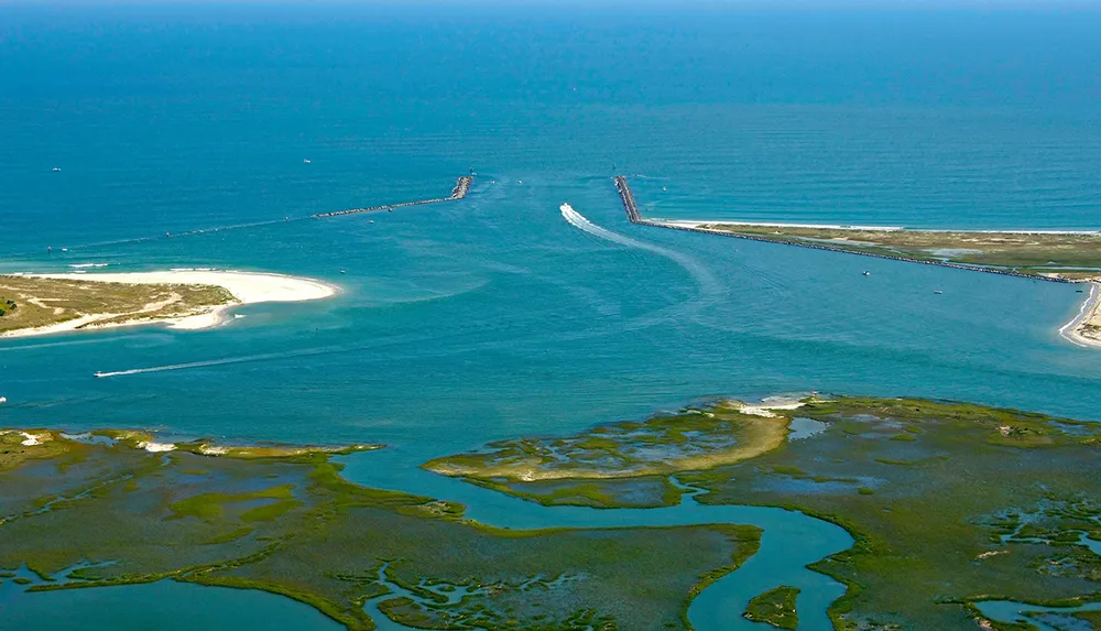 This is an aerial view of a coastal inlet with protective breakwaters showing boats navigating the channel and surrounding wetlands