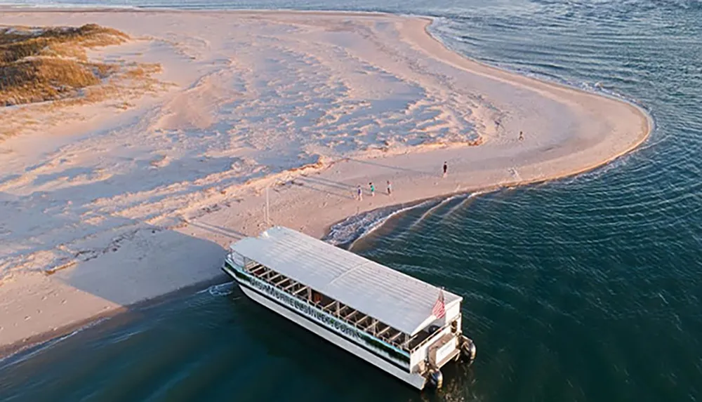 A boat is moored near a narrow strip of sand with people walking around surrounded by water on both sides during what appears to be low tide