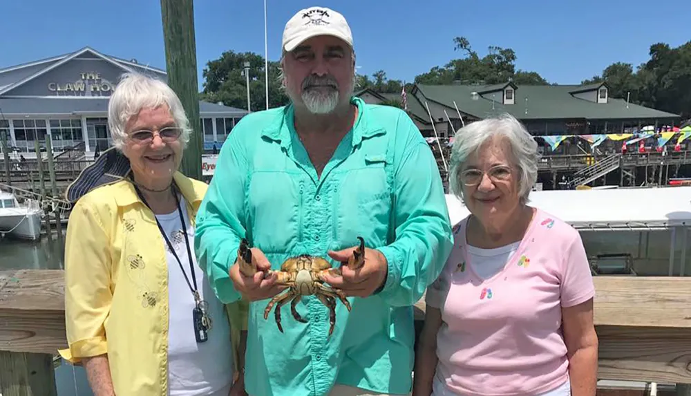 A man in a teal shirt is holding a crab with two smiling women on either side against the backdrop of a waterfront restaurant called The Claw House