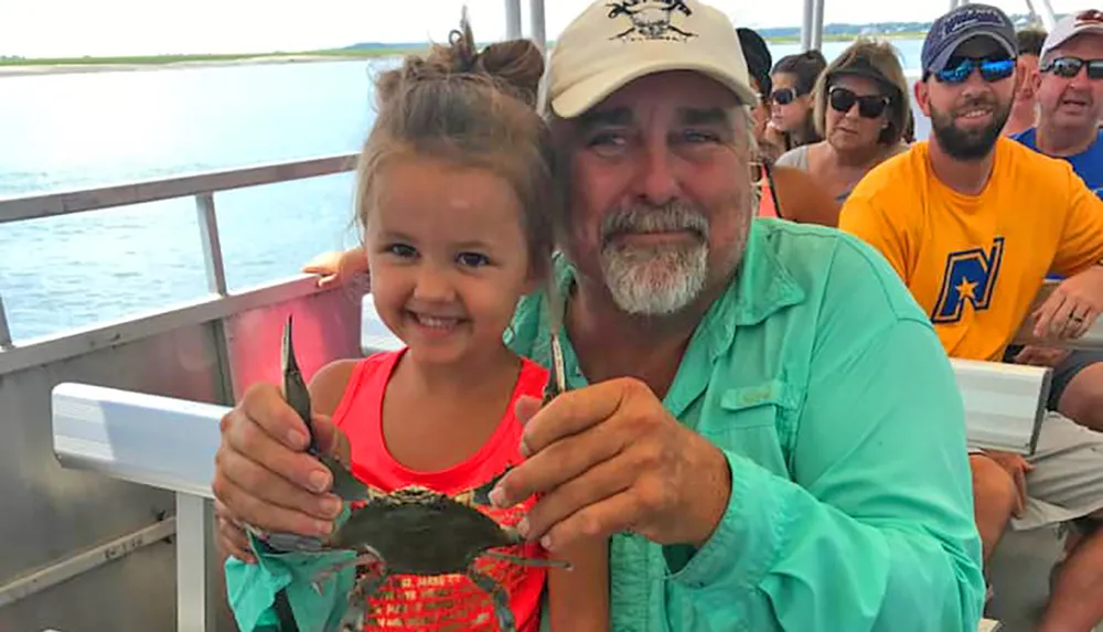 A smiling girl and an older man are showing a crab to the camera on a boat with other passengers in the background