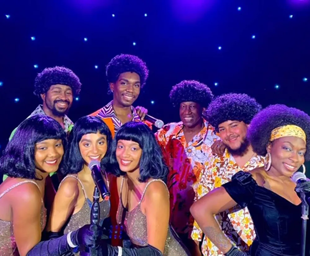 A group of performers is wearing retro 1970s-style outfits with afros and colorful clothing likely representing a music group from that era posing together on stage with a starry background