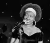 A woman with a microphone is smiling on stage possibly during a performance or speech with a backdrop of blue lights