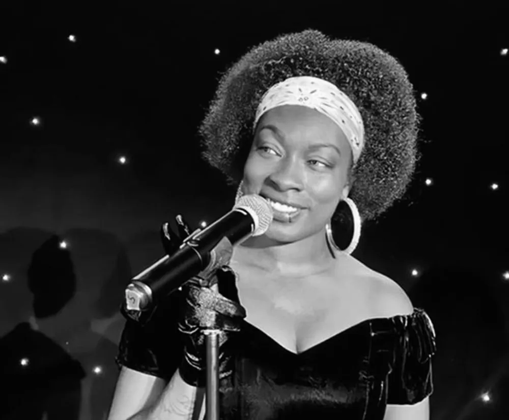 A woman with a headband and hoop earrings smiles while singing into a microphone on a stage with a starry backdrop