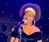 A woman with a microphone is smiling on stage possibly during a performance or speech with a backdrop of blue lights