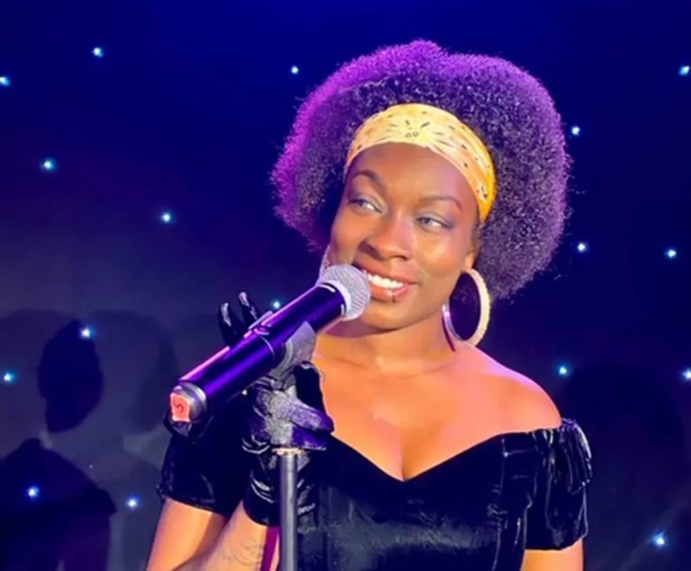 A woman with a microphone is smiling on stage possibly during a performance or speech with a backdrop of blue lights