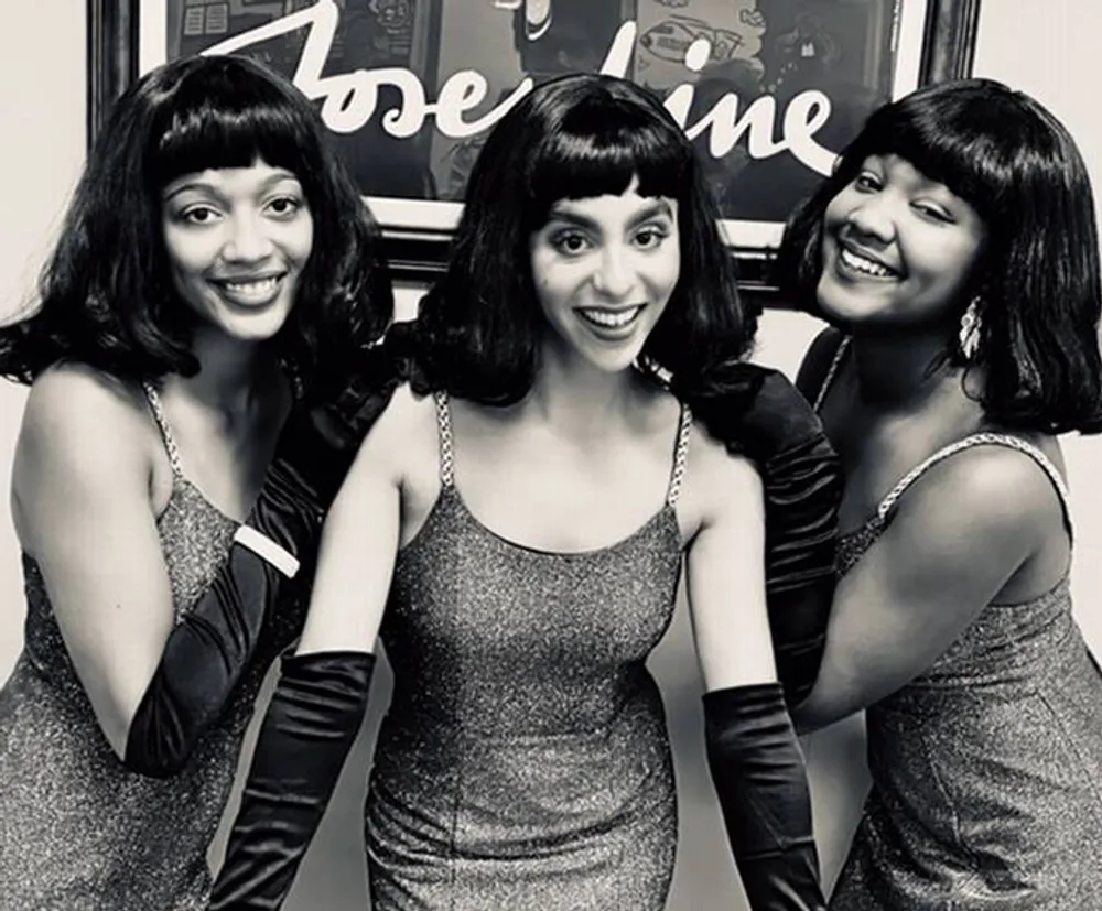 Three women dressed in matching silver dresses and black gloves wearing similar hairstyles pose together with smiles giving a vintage or retro vibe in a black-and-white photograph