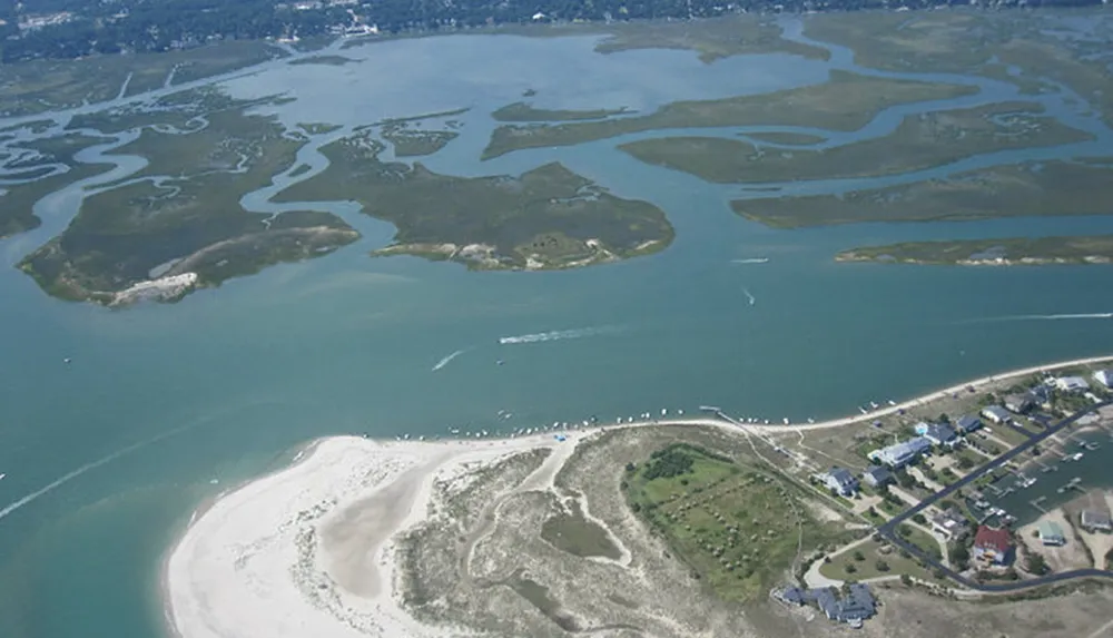 This is an aerial photo of a coastal area featuring meandering water channels green wetlands a stretch of beach and a small residential community
