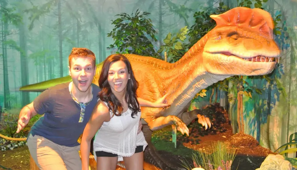 A man and a woman are posing playfully in front of a model of a Tyrannosaurus rex in a forest-themed display