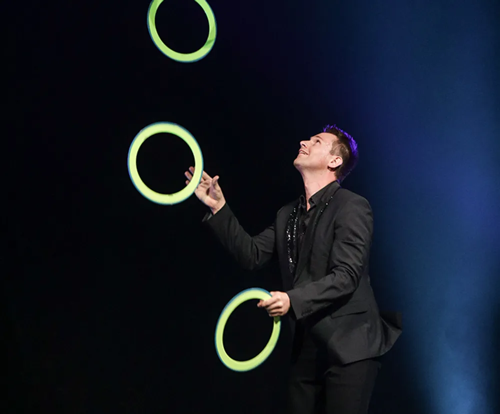 A performer in a black outfit is juggling multiple glowing green rings against a dark background