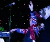 A performer in an extravagant costume with a feather boa sings into a microphone under colorful stage lighting with the word BOOM graphically overlaid on their hat