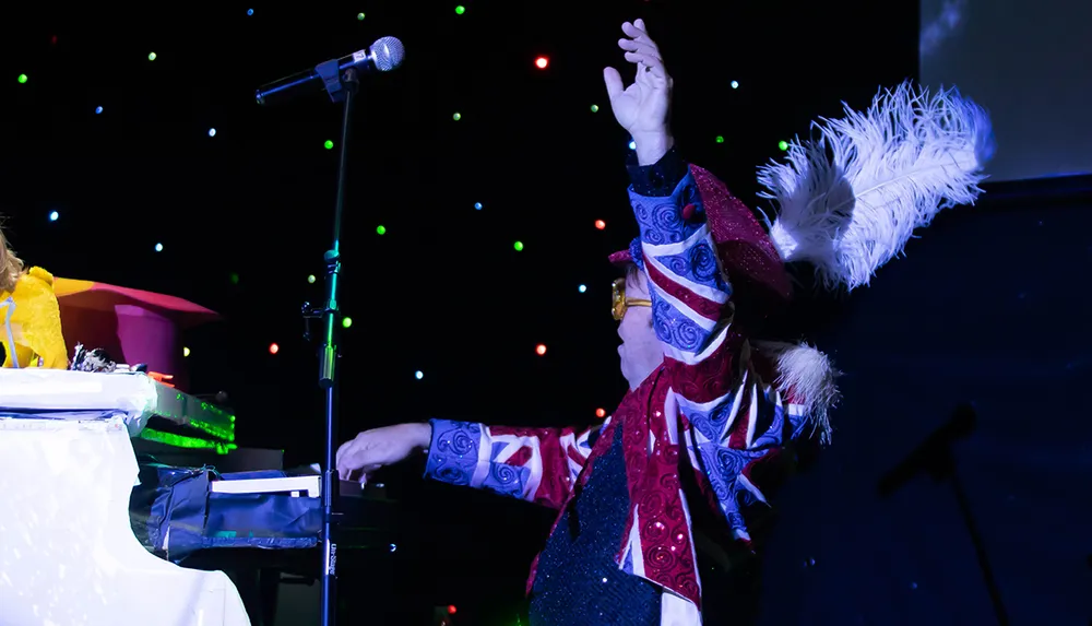 A flamboyantly dressed performer is passionately playing a piano on a stage with a colorful backdrop of lights