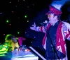 A performer in an extravagant costume with a feather boa sings into a microphone under colorful stage lighting with the word BOOM graphically overlaid on their hat
