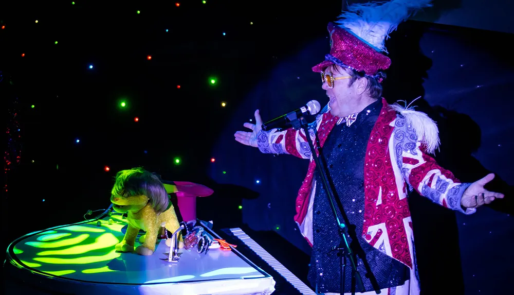 A performer dressed in a flamboyant costume and top hat is singing into a microphone while another person in a lion costume plays a keyboard on a stage with colorful lights scattered in the background