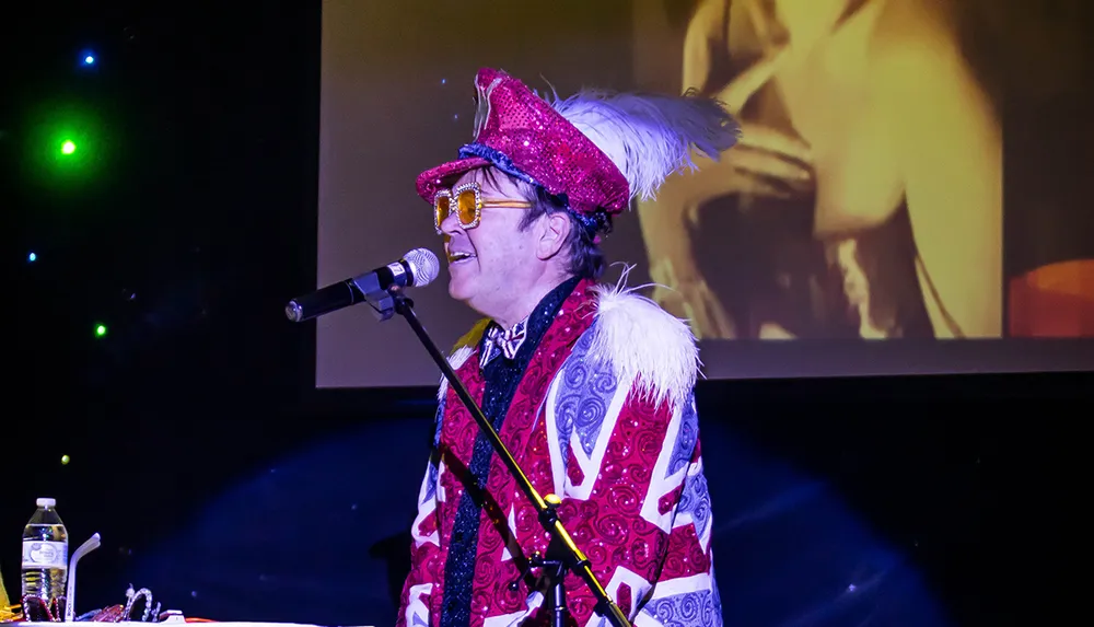 A person in a flamboyant and colorful outfit including a sequined hat and glasses is performing on stage with a microphone