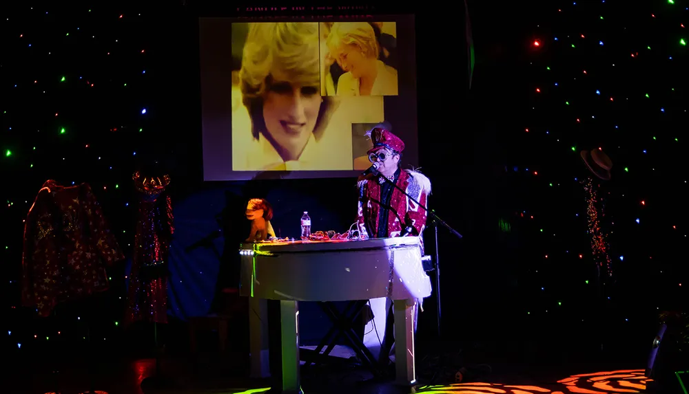 A person in an elaborate costume and glasses is playing a white keyboard on a stage adorned with colorful lights and a video projection in the background