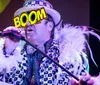 A performer in an extravagant costume with a feather boa sings into a microphone under colorful stage lighting with the word BOOM graphically overlaid on their hat