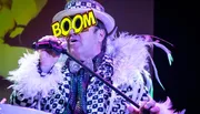 A performer in an extravagant costume with a feather boa sings into a microphone under colorful stage lighting, with the word