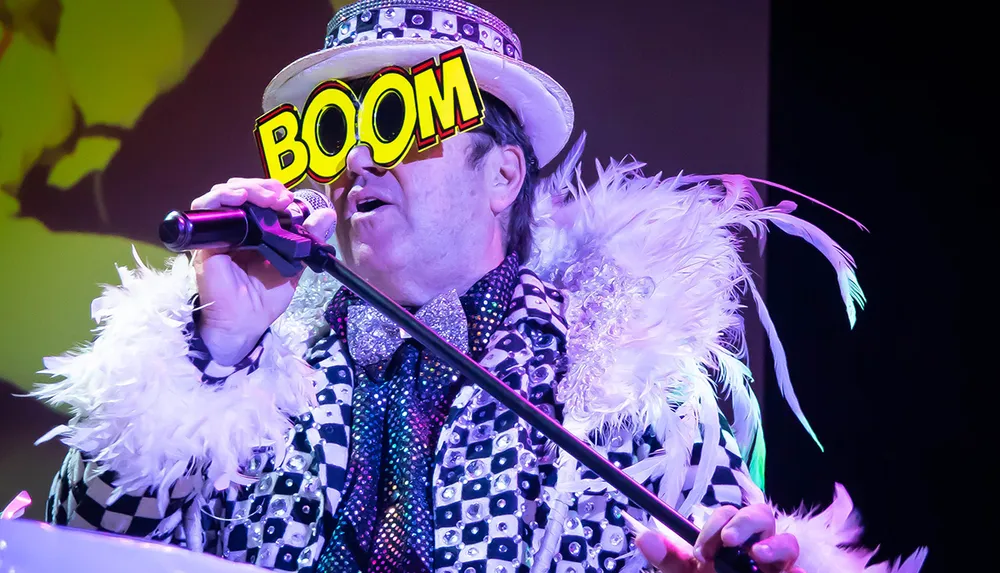 A performer in an extravagant costume with a feather boa sings into a microphone under colorful stage lighting with the word BOOM graphically overlaid on their hat