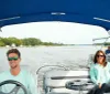 Two individuals are enjoying a ride on a blue and white pontoon boat with a blue sunshade on a calm river surrounded by trees
