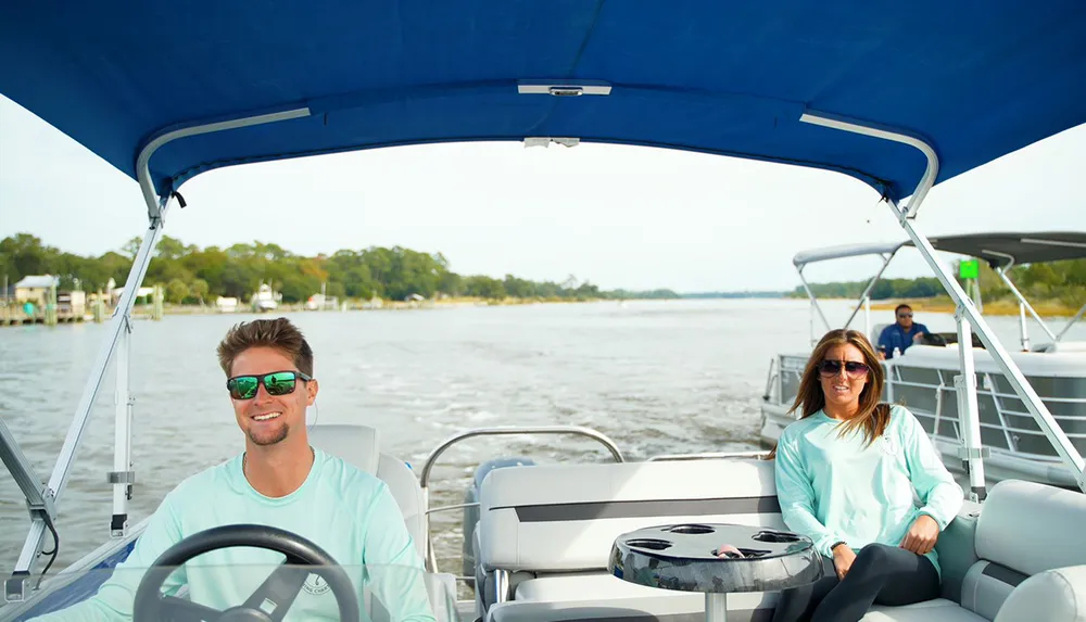 A man and a woman are enjoying a sunny day on a boat with the man at the helm and the woman seated at a table both smiling and wearing sunglasses