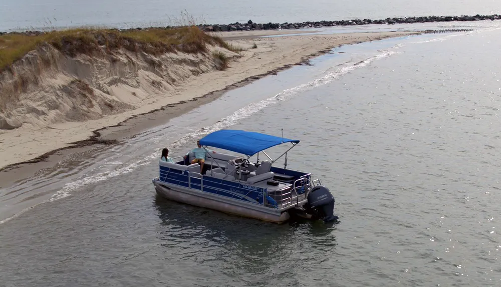 A pontoon boat with two occupants is navigating close to the sandy shore near a rocky inlet