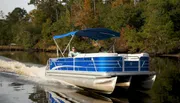 Two individuals are enjoying a ride on a blue and white pontoon boat with a blue sunshade on a calm river surrounded by trees.