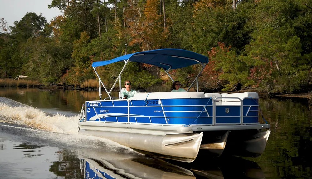 Two individuals are enjoying a ride on a blue and white pontoon boat with a blue sunshade on a calm river surrounded by trees