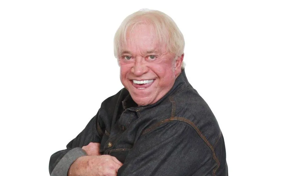 A man with blond hair is smiling broadly at the camera wearing a dark button-up shirt with his arms crossed