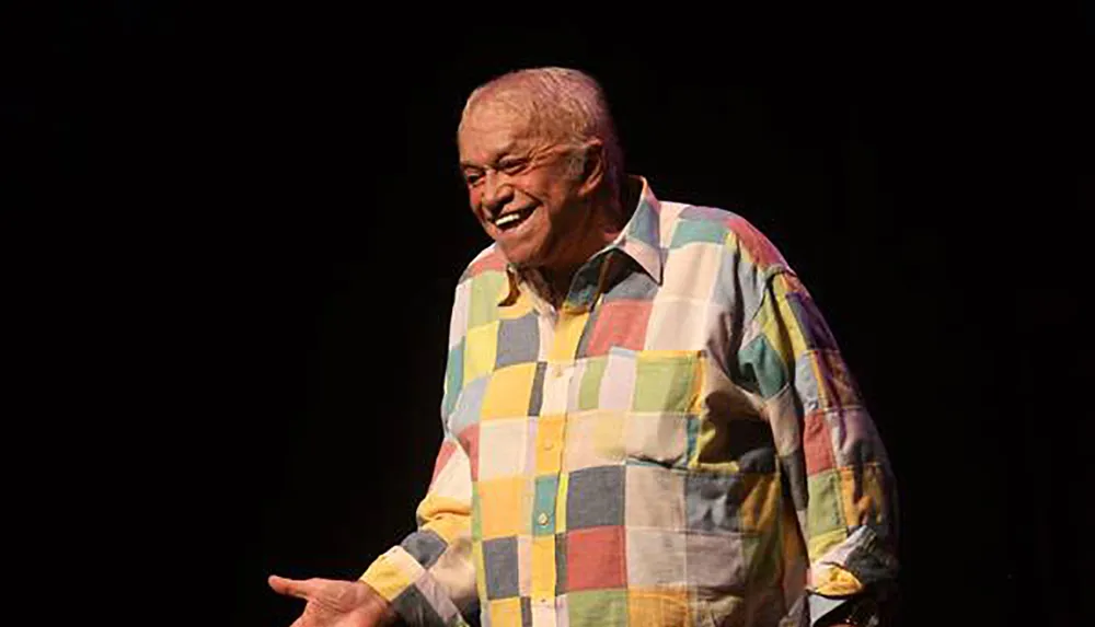 A man is standing on stage wearing a colorful checkered shirt smiling broadly as if engaging with an audience